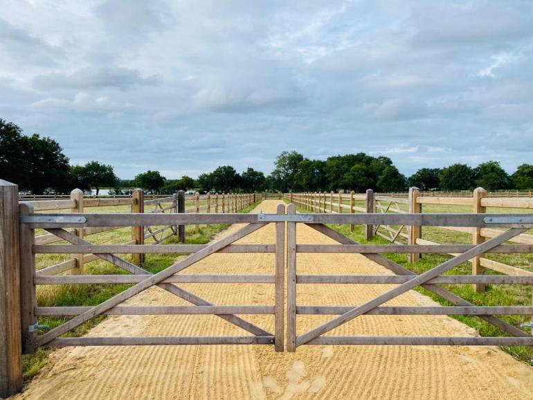 A wooden gate on a sand path with two fields next to it fenced with wooden fences.