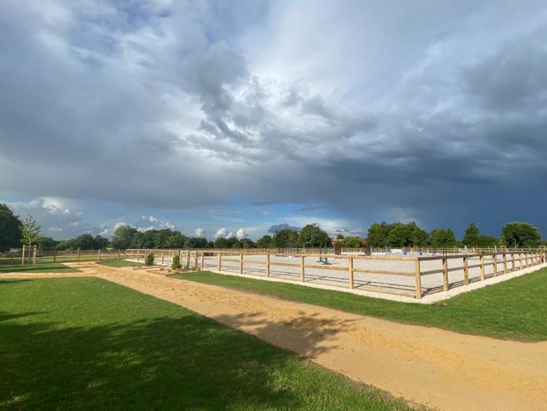 A sand path with grass around it and a riding arena on the back that has wooden fences around it.