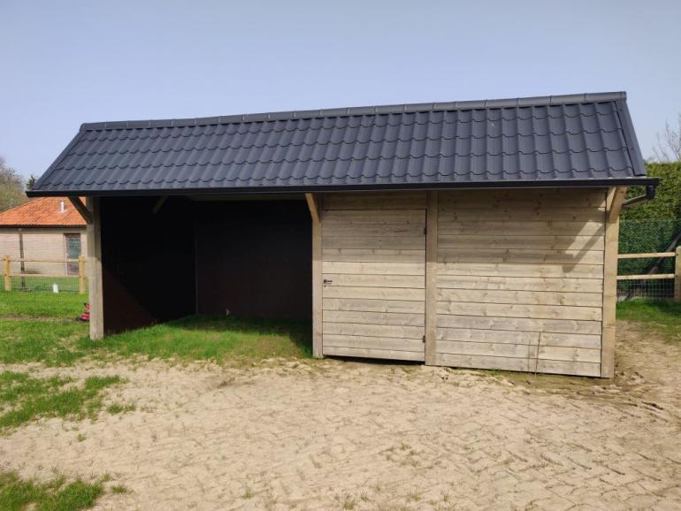Een houten schuilstal voor buitendieren met een opbergplaats, een deur en zwarte dakpannen