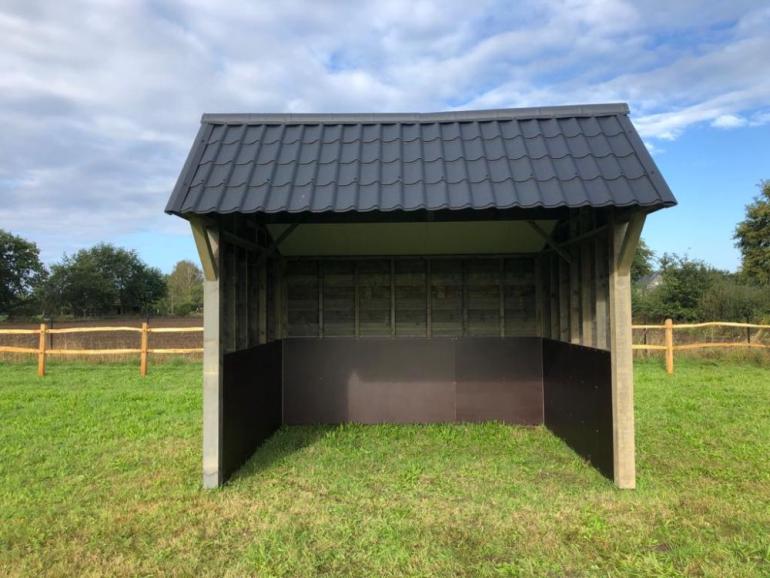 Een houten schuilstal voor paarden en buitendieren met zwarte dakpannen op een veld met houten omheining