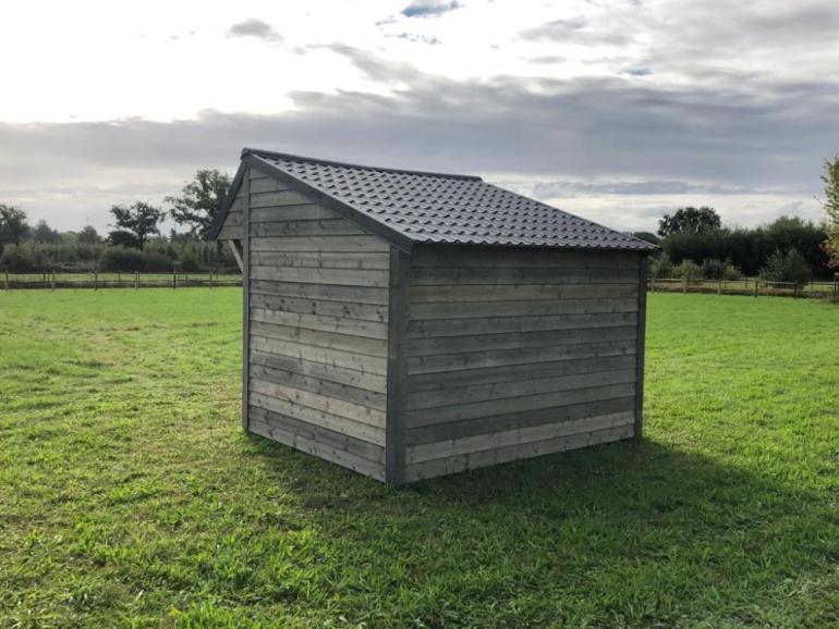 Een houten schuilstal voor buitendieren met zwarte dakpannen op een veld