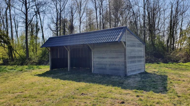 Een dubbele houten paardenschuilplaats met een opslagruimte eraan vast en zwarte dakpannen