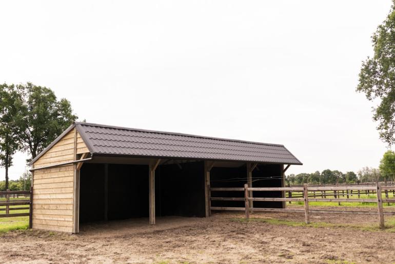 Twee dubbele houten schuilstallen voor paarden met zwarte dakpannen op een veld met houten omheining met drie sporen