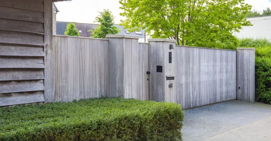 A sliding wooden gate for residential properties with vertical boards next next to a door with an intercom system